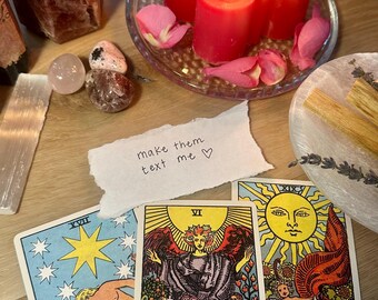 May include: A collection of spiritual items on a wooden surface. Includes lit red candles, tarot cards, crystals, palo santo sticks, and a handwritten note that says "make them text me". The tarot cards feature illustrations and the words "The Lovers" and "The Sun".
