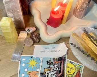 May include: An arrangement of crystals, tarot cards, candles, and palo santo sticks. The tarot cards include "The Star," "Queen of Cups," and "The Sun." A handwritten note reads "self love and confidence."