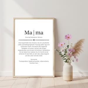 May include: A framed print with the German word "Mama" in a stylish font. The print includes a definition and synonyms in German. A vase with pink and white flowers and decorative grass sits to the right of the frame.