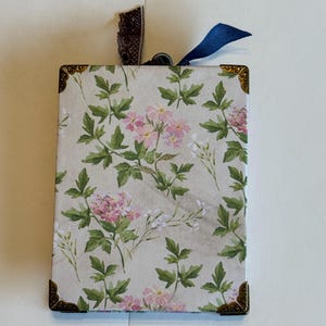 Peut inclure: Boîte rectangulaire à motifs floraux avec des fleurs roses et des feuilles vertes sur fond crème. La boîte a des coins décoratifs, un ruban bleu foncé et un ruban en dentelle marron.