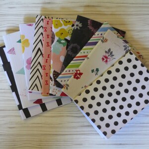 May include: A stack of eight small notebooks with different patterned covers. The covers feature geometric patterns, floral designs, and polka dots in various colors, including black, white, pink, red, and green.