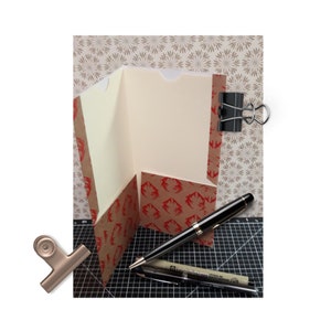 May include: A brown paper folder with a red and brown patterned design. The folder is folded in half and has a white interior. A black pen and a silver pen are lying on a black cutting mat.