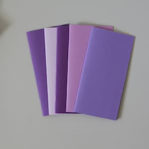 May include: A collection of six blank notebooks in shades of purple, ranging from light lavender to deep violet. The notebooks are arranged in a fan shape, showcasing their different hues. The paper appears smooth and the covers are a solid color.