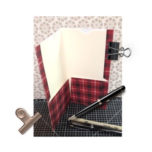 May include: A red and black plaid folder with a white interior and a flap for holding papers. The folder is closed and has a silver binder clip attached to the top. A black pen is lying on a black cutting mat.