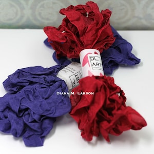 May include: Two spools of ruffled fabric ribbon, one red and one purple, with a white label that reads "DL ART" and "DIANA M. LARSON".