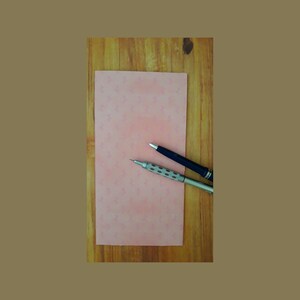 May include: A pink sheet of paper with a subtle pattern lies on a wooden surface. Two writing utensils, a black pen and a silver mechanical pencil, are placed on the paper.