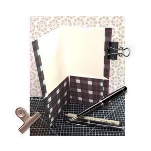 May include: A black and white plaid patterned paper notebook with a cream colored interior. The notebook is closed and held together with a black binder clip. Two pens are lying on a black and white grid surface.