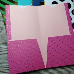 May include: A bright pink two-pocket folder featuring a pattern of small gold dots. The interior of the folder is a solid light pink color, and the two bottom pockets have semi-circular cutouts at the top edges for easy access to documents.