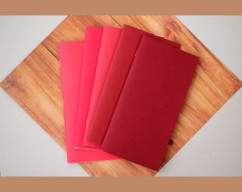 May include: Five red notebooks of varying shades, stacked on a wooden surface.