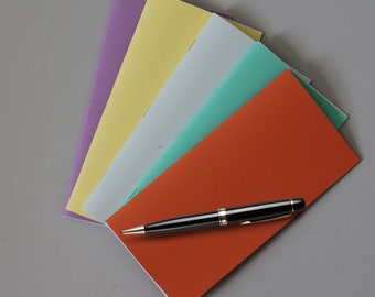 May include: A collection of colorful, rectangular notebooks in shades of purple, yellow, light blue, teal, and orange. A black and gold pen rests on top of the orange notebook, suggesting a stationery or office supply theme.