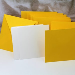 May include: A stack of yellow cardstock envelopes and blank white cards. The envelopes are folded and stacked on top of each other. The cards are also stacked on top of each other.