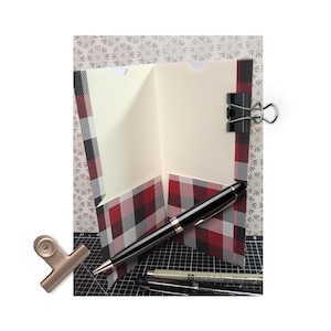 May include: A red and black plaid card with a white interior. The card is folded in half and has a pocket on the inside. A black pen is lying on top of the card.