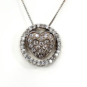 May include: A silver necklace with a heart-shaped pendant. The pendant features a heart-shaped design within a circle, both encrusted with small, clear gemstones. The chain is a delicate, silver link style.