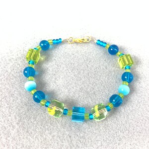 May include: A blue and green beaded bracelet with a gold clasp. The bracelet features round and square beads in shades of blue and green.