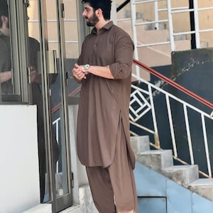May include: A brown, long-sleeved tunic and matching trousers. The outfit has a collared neckline and side slits on the tunic. The person is wearing black sandals and a silver watch. The outfit is a traditional style.