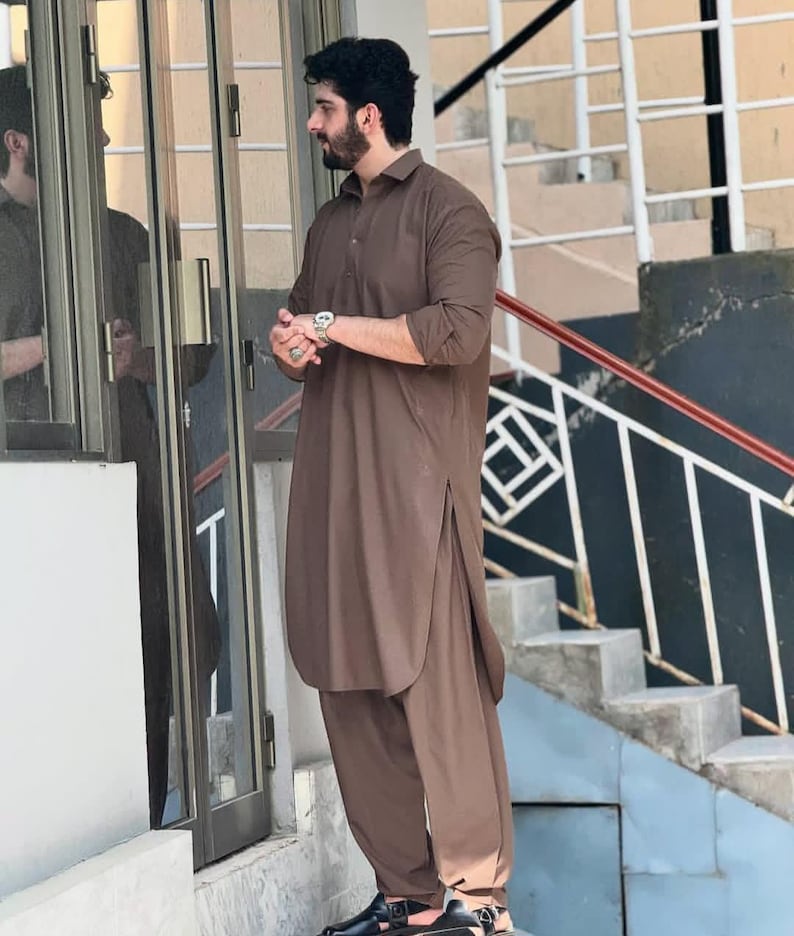 May include: A brown, long-sleeved tunic and matching trousers. The outfit has a collared neckline and side slits on the tunic. The person is wearing black sandals and a silver watch. The outfit is a traditional style.