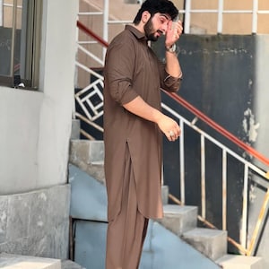 May include: A brown salwar kameez outfit. The long-sleeved shirt has a collared neckline and side slits. The matching pants are loose-fitting. The outfit is worn by a person standing on a set of stairs.