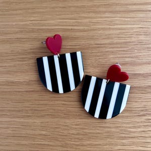 May include: A pair of earrings featuring a black and white striped semi-circle design with a small red heart accent. The earrings are set against a wooden background.