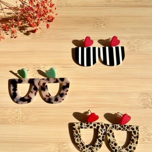 May include: Three pairs of U-shaped dangle earrings. One pair features black and white vertical stripes with red heart studs. Another pair has a tortoiseshell pattern with mint green heart studs. The third pair shows a spotted pattern with red heart studs.