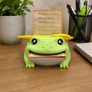 May include: A light green frog-shaped desk organizer with a wide mouth holding a stack of orange sticky notes. A yellow pen rests horizontally across the frog's head. The frog has large black and white eyes and a textured surface finish.
