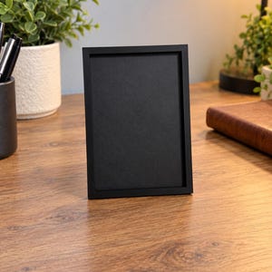 May include: A rectangular picture frame with a thin, matte black border and a solid black interior panel. The frame is oriented vertically, featuring clean, straight edges and a flat, uniform surface finish, designed to hold a single photograph or print.
