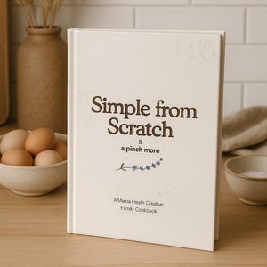 May include: A hardcover cookbook titled "Simple from Scratch & a pinch more" stands on a wooden surface. The cover is off-white with brown text and a sprig of purple flowers. The cookbook is a family cookbook by A Mama Heath Creative.