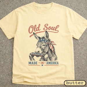 May include: Cream-coloured t-shirt with a detailed illustration of a donkey playing a harmonica, wearing a bandana with a star-spangled design. The text "Old Soul" arches above the donkey, with "MADE IN AMERICA" below.
