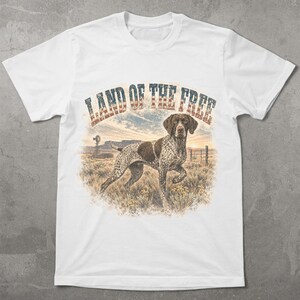 May include: A white short-sleeve t-shirt featuring a graphic of a German Shorthaired Pointer standing in a field at sunset. The text LAND OF THE FREE is printed in a distressed American flag pattern above the dog, with a windmill in the background.