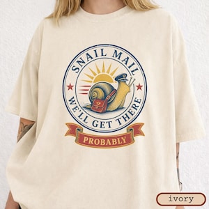 May include: An ivory t-shirt featuring a circular graphic of a snail wearing a mail carrier hat and a satchel. The text reads SNAIL MAIL WE'LL GET THERE PROBABLY. The design includes a yellow sun, red stars, and a red banner on a cream background.