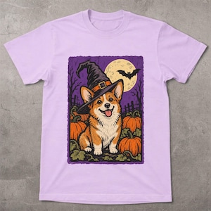 May include: A light purple t-shirt featuring a graphic illustration of a Corgi dog wearing a black witch hat. The scene includes orange pumpkins, a large yellow full moon, and a black bat silhouette against a dark purple background.
