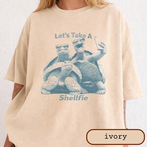 Puede incluir: Camiseta color marfil con un gráfico azul claro de tres tortugas con gafas de sol y el texto "Let's Take A Shellfie". La camiseta tiene un corte holgado.