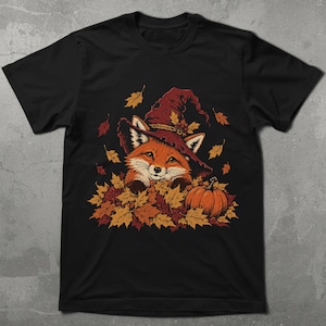 Puede incluir: Una camiseta negra de manga corta con un estampado gráfico de un zorro con un sombrero marrón puntiagudo, metido entre un montón de hojas de otoño naranjas y amarillas con una pequeña calabaza al lado y hojas cayendo alrededor.