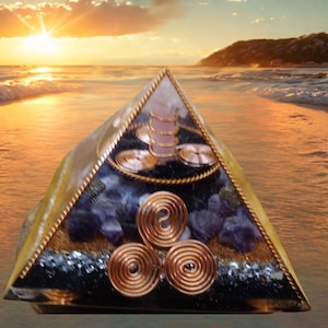 May include: An orgone energy pyramid with a gold frame, amethyst stones, and copper coils. The pyramid has a central crystal and sits against a beach sunset backdrop. The pyramid's dimensions are approximately 10 cm.