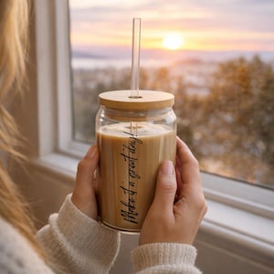 Puede incluir: Un vaso de vidrio transparente lleno de una bebida marrón claro, con una tapa de bambú y una pajita transparente. Las palabras "Make it a great day!" están impresas en el vaso. El vaso se sostiene frente a una ventana con una vista del atardecer.