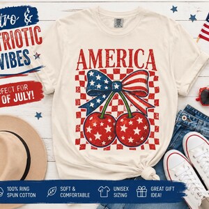 Peut inclure: T-shirt crème avec un motif de cerises patriotiques. Le mot "AMERICA" est imprimé au-dessus d'un nœud et de deux cerises avec des étoiles rouges et blanches. Le t-shirt porte également le texte "Retro & Patriotic Vibes" et "Perfect for 4th of July!"