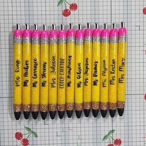 May include: A collection of glitter pens designed to resemble pencils. Each pen is yellow with a silver glitter band, a pink glitter top, and a black tip. The pens are personalized with names in black script, perfect for teachers.