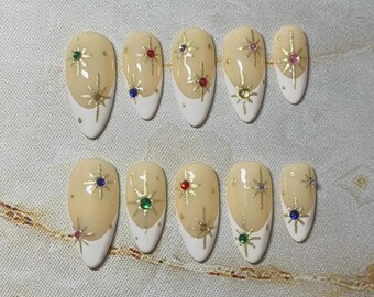 May include: A set of ten almond-shaped press-on nails featuring a nude base with white French tips. Each nail is decorated with gold starburst patterns and small, colorful embedded rhinestones in shades of red, green, blue, and pink.