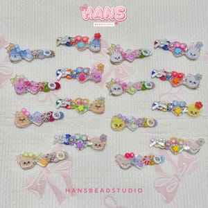 Pode incluir: Uma coleção de presilhas de cabelo coloridas com vários designs de personagens, incluindo coelhos, gatos e outras figuras fofas. As presilhas apresentam uma mistura de contas, corações e detalhes florais, tudo em um fundo claro. A marca "HANS BEADSTUDIO" é visível.