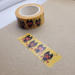May include: A roll of orange washi tape with a repeating pattern of black and red cartoon characters. A small piece of tape with the same design is shown below the roll. The tape is on a brown surface.