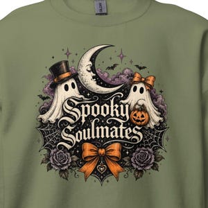 Könnte beinhalten: Ein olivgrünes Sweatshirt mit Rundhalsausschnitt und einem Grafikdruck von zwei Geistern, einer Mondsichel, einem Kürbis und Rosen. Der Text "Spooky Soulmates" ist mittig platziert, ergänzt durch eine große orangefarbene Schleife, Spinnweben und Fledermäuse.