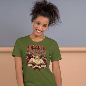 May include: An olive green short-sleeve t-shirt featuring a graphic print of an open book with a diamond ring resting on the pages, surrounded by red roses and greenery. The text reads Plot Twist: I'm Engaged! with smaller text below the book.