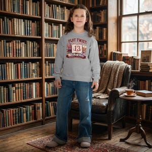 May include: A light gray long-sleeve sweatshirt featuring a graphic print that says PLOT TWIST: She Turns 13, with illustrations of stacked books, an open book with the number 13, a coffee cup, and a pair of glasses, paired with blue denim jeans.