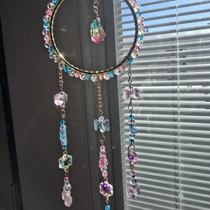 May include: A decorative suncatcher with a gold-colored metal ring, adorned with colorful beads in shades of pink, blue, and purple. Dangling from the ring are strands of beads and crystal-like shapes, creating a shimmering effect. The suncatcher is hanging near a window.