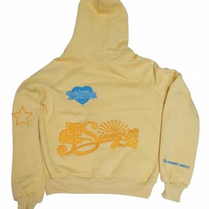 May include: A pale yellow hoodie with a hood and long sleeves. The back features embroidered text and graphics, including a blue heart with "Forever" and "BES" with a sunburst design. A star is on the left sleeve, and text on the right sleeve.