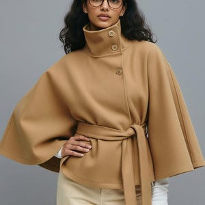 Puede incluir: Un abrigo tipo capa de color camel con cuello alto y cierre de botones. El abrigo tiene un cinturón a juego atado a la cintura. La modelo lleva gafas y una camisa blanca de manga larga. El abrigo está hecho de una tela suave.