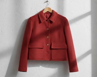 May include: A red jacket with a classic design, featuring a collared neckline, two front pockets, and gold-colored buttons. The jacket is hanging on a wooden hanger against a white wall.