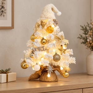 Op de afbeelding: Een witte, besneeuwde kunstkerstboom voor op tafel met een bijpassende gebreide muts als piek. De boom is versierd met gouden kerstballen, strikken, een ster en een houten label met een sneeuwvlok. De voet is omwikkeld met jute en verlicht.