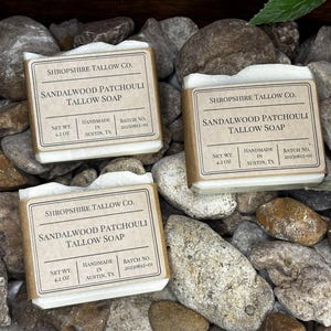 May include: Three bars of handmade soap with labels that read "Shropshire Tallow Co. Sandalwood Patchouli Tallow Soap." Each bar weighs 4.2 oz. The soaps rest on a bed of gray and brown stones.
