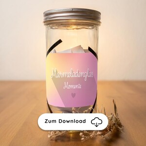 May include: A clear glass jar with a silver lid, containing white slips of paper. A pink and yellow label with the words "Marmeladenglas Momente" is wrapped around the jar. The jar is on a wooden surface.