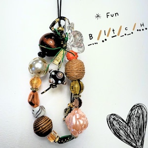 Può includere: Un braccialetto con ciondoli di perline con una varietà di perline colorate, tra cui perle, vetro ed elementi strutturati. La parola "Fun" è scritta sopra il braccialetto e la parola "Bitch" è scritta in codice Morse. Un cuore nero è disegnato nell'angolo.